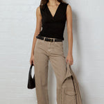 A woman stands against a white brick wall wearing a black sleeveless wrap top, beige wide-leg cargo pants, and black patterned sandals, while holding a black handbag and a beige jacket.