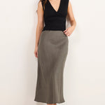 A woman modeling a sleeveless black wrap-style top and a long, straight, knee-length olive green skirt, paired with black flip-flop sandals, against a plain off-white studio background.