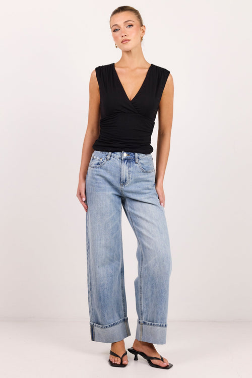 A model stands against a plain white studio background wearing a black sleeveless wrap-style top, light-wash wide-leg jeans, and black sandals.