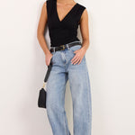 A woman modeling a black sleeveless V-neck top, light-wash wide-leg jeans, black sandals, and sunglasses, holding a small black handbag against a plain white studio background.