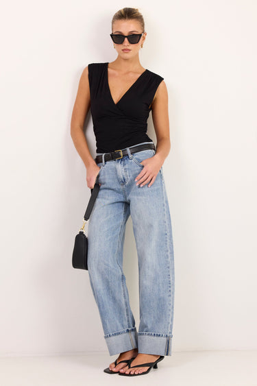 A woman modeling a black sleeveless V-neck top, light-wash wide-leg jeans, black sandals, and sunglasses, holding a small black handbag against a plain white studio background.