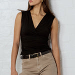 A woman stands against a white brick wall wearing a black sleeveless wrap top, beige cropped pants, a black belt, black-and-brown patterned flat shoes, sunglasses, and holding a black clutch and a beige duffel bag.