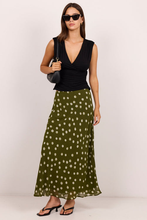 A woman modeling a sleeveless black top and a flowing olive green midi skirt with white polka dots, styled with black sandals, sunglasses, and a black shoulder bag against a plain light background.