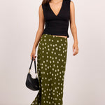 A woman modeling a sleeveless black wrap-style top and a flowing olive green skirt with white polka dots, holding a black handbag against a plain light gray studio background.