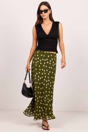 A woman modeling a sleeveless black wrap-style top and a flowing olive green skirt with white polka dots, holding a black handbag against a plain light gray studio background.