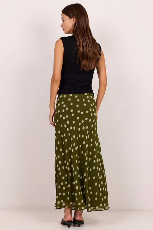 A woman is shown from the back wearing a sleeveless black top and a long olive green skirt with a white polka dot pattern, standing against a plain off-white studio background.