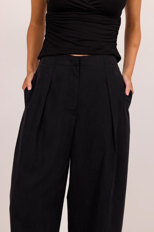 A close-up of a person wearing black wide-leg pleated trousers with side pockets, paired with a black ruched top, against a plain light background.
