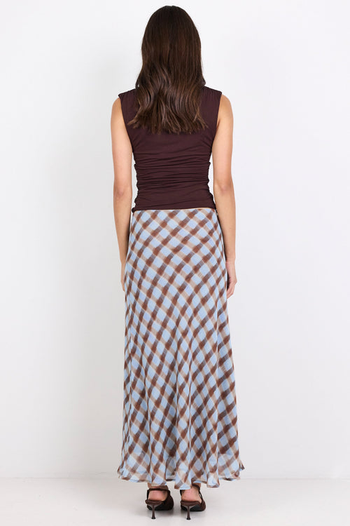 A woman is shown from behind wearing a sleeveless dark brown top and a long skirt with a brown, beige, and light blue gingham pattern, standing against a plain white studio background.