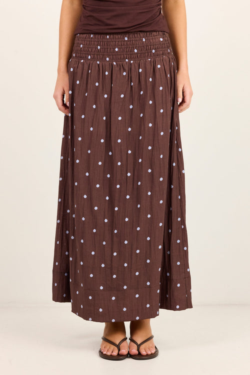 A model wearing a brown pleated maxi skirt with a white polka dot pattern, paired with a matching brown top and brown sandals, against a plain off-white studio background.