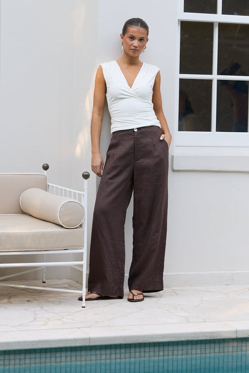 A woman stands outdoors by a pool wearing a white sleeveless wrap-style top and dark brown wide-leg pants, with a white building and window behind her.