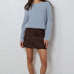 A woman stands against a white brick wall wearing a light blue knit sweater, a dark brown mini skirt, and dark brown slip-on shoes.