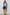 A woman stands against a white brick wall wearing a light blue knit sweater, a dark brown mini skirt, and dark brown slip-on shoes.
