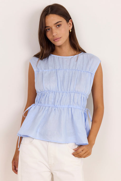 A woman modeling a light blue sleeveless peplum top with a gathered elastic waist and drawstring ties, paired with white pants, against a plain off-white background.