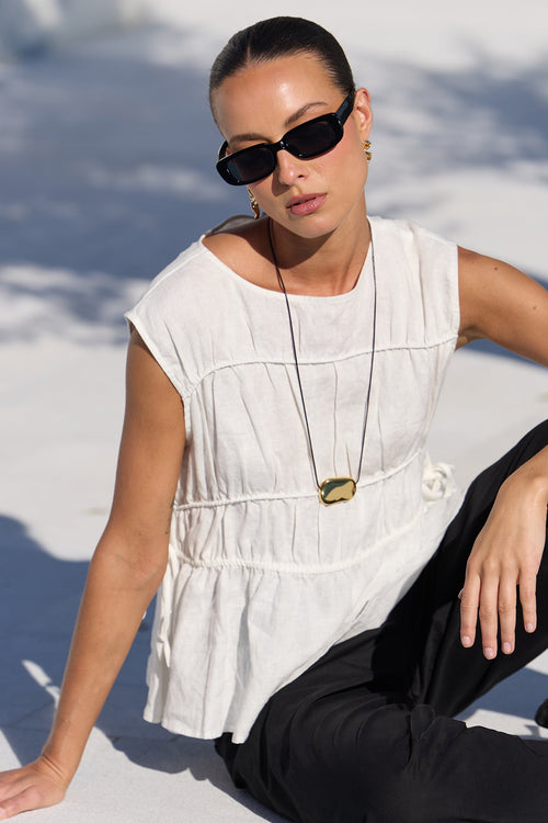 A woman wearing a white sleeveless peplum top, black pants, and black sunglasses sits on a concrete poolside ledge in bright sunlight.