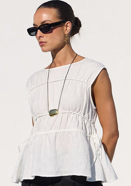 A woman stands by a pool wearing a white sleeveless peplum top, black cropped wide-leg pants, and black sandals in a bright white architectural setting.