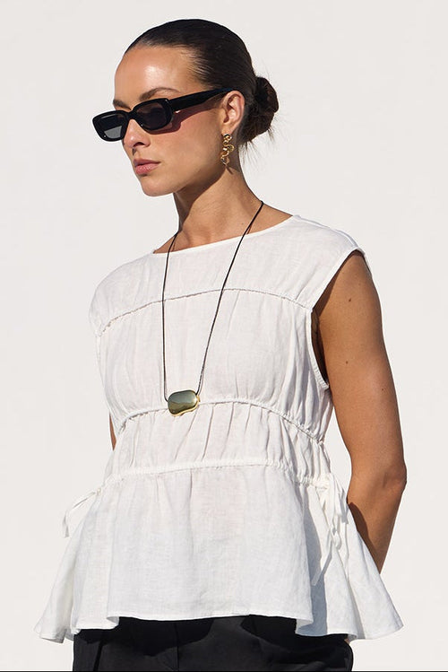 A woman stands by a pool wearing a white sleeveless peplum top, black cropped wide-leg pants, and black sandals in a bright white architectural setting.