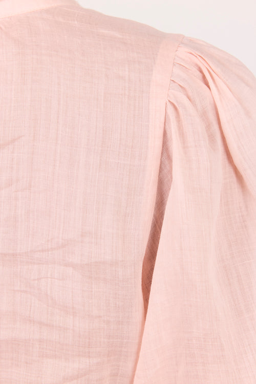 Close-up of a light pink textured fabric garment showing a shoulder seam and pleated detail against a plain light background.