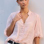 A woman modeling a light pink linen button-up blouse with white pants and a black belt, standing against a white wall.