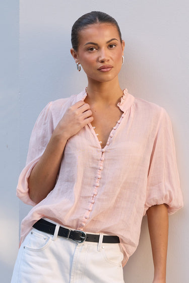 A woman modeling a light pink linen button-up blouse with white pants and a black belt, standing against a white wall.