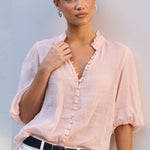 A woman wearing a light pink short-sleeve button-front blouse with ruffled trim, tucked into white pants with a black belt, standing against a white wall.