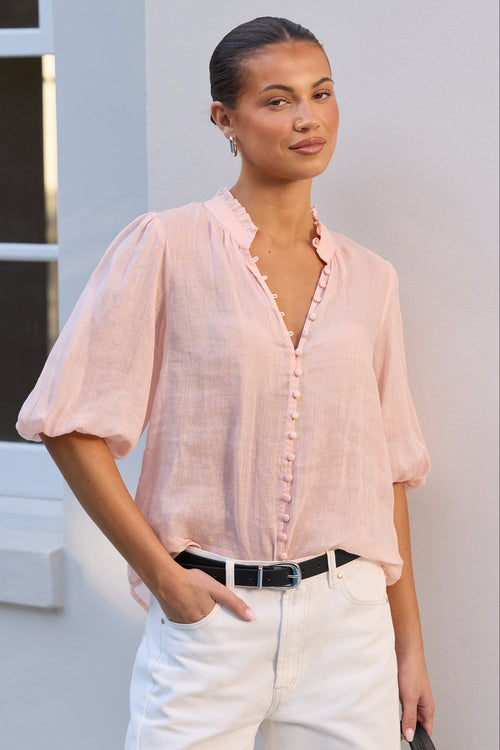 A woman wearing a light pink short-sleeve button-up blouse with ruffled trim, tucked into white trousers with a black belt, standing against a light-colored wall.