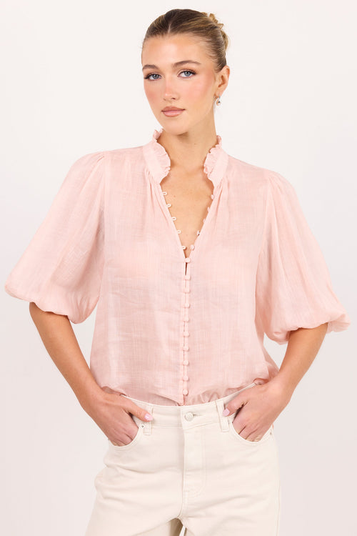 A woman modeling a light pink short-sleeve blouse with a ruffled high collar and button-front placket, paired with off-white pants, against a plain light background.