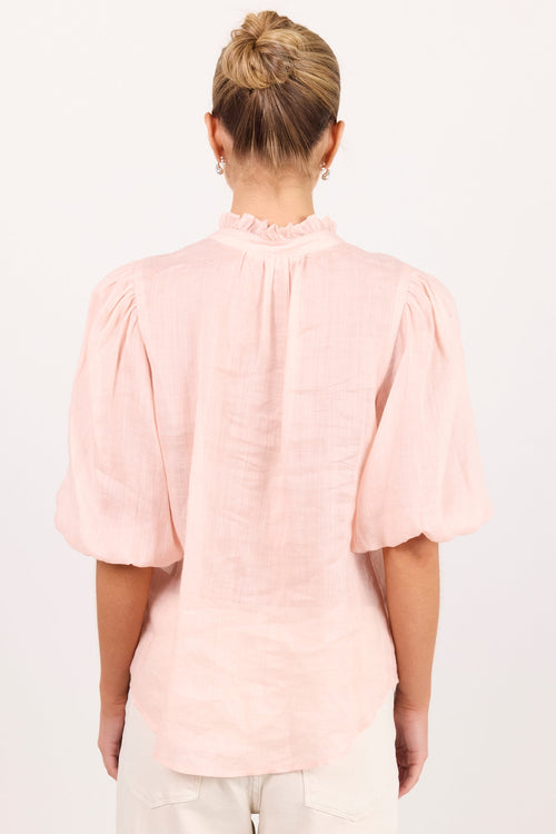 A woman viewed from the back wearing a light pink linen blouse with short puff sleeves and a ruffled mock neck, paired with off-white pants, against a plain white background.