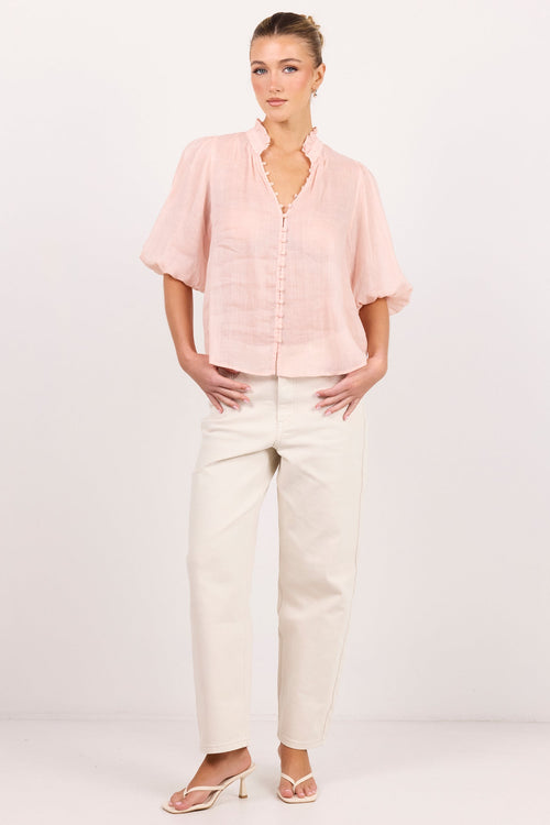 A woman modeling a light pink short-sleeve button-up blouse with a ruffled collar and front buttons, paired with off-white trousers and beige heeled sandals, against a plain white studio background.