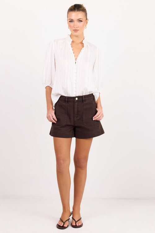 A woman modeling a white long-sleeve button-up shirt and dark brown shorts against a plain white studio background.