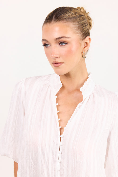 A woman with blonde hair in a bun wearing a white crinkled blouse with a ruffled collar and button-front closure, photographed against a plain white background.