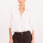 A woman modeling a white long-sleeve button-up blouse with a split neckline and dark brown shorts against a plain white studio background.