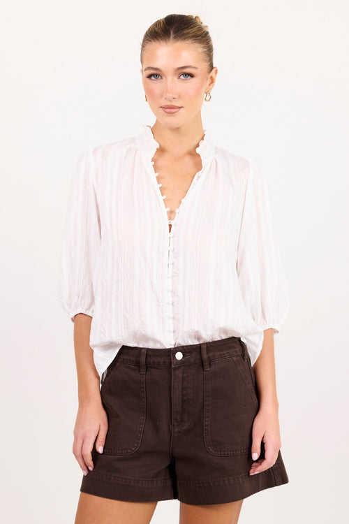 A woman modeling a white long-sleeve button-up blouse with a split neckline and dark brown shorts against a plain white studio background.