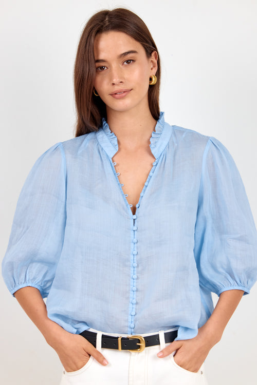 A woman wearing a light blue blouse with puff sleeves, a ruffled collar, and a ruffled front placket, paired with white pants and a black belt, against a light gray studio background.