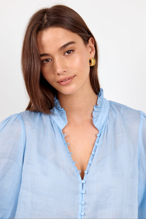 A woman with shoulder-length brown hair wearing a light blue ruffled blouse and gold hoop earrings, photographed against a plain light gray studio background.