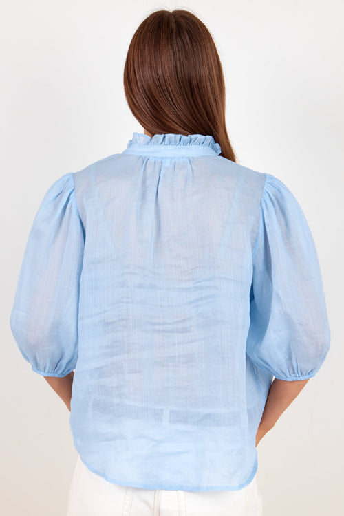 A woman viewed from the back wearing a light blue sheer puff-sleeve blouse with a ruffled collar, paired with white pants, against a plain off-white background.
col_pattern