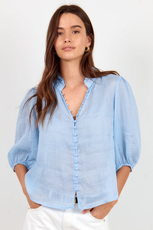 A woman modeling a light blue long-sleeve button-up blouse with puffed shoulders, paired with white wide-leg pants and black sandals, standing against a plain white studio background.