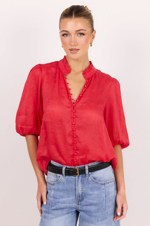 A woman modeling a red short-sleeve blouse with a ruffled V-neckline and button front, paired with light blue jeans and a black belt, against a plain light background.
