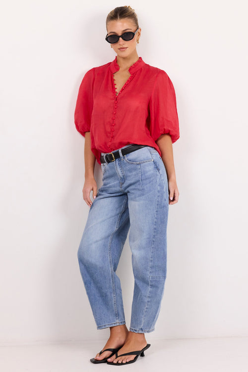 A woman modeling a vibrant red short-sleeve blouse with puffed shoulders and a ruffled collar, paired with light-wash blue jeans, a black belt, and black sandals against a plain white studio background.
