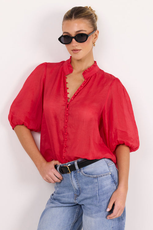 A woman modeling a red short-sleeve blouse with a ruffled collar and button front, paired with light blue jeans and black sunglasses, against a plain white background.