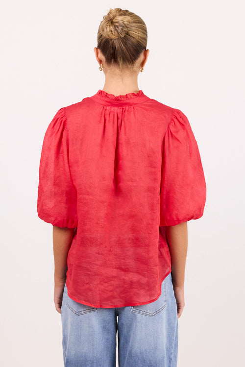 A woman viewed from behind wearing a red short puff-sleeve blouse and light blue jeans, standing against a plain white studio background.