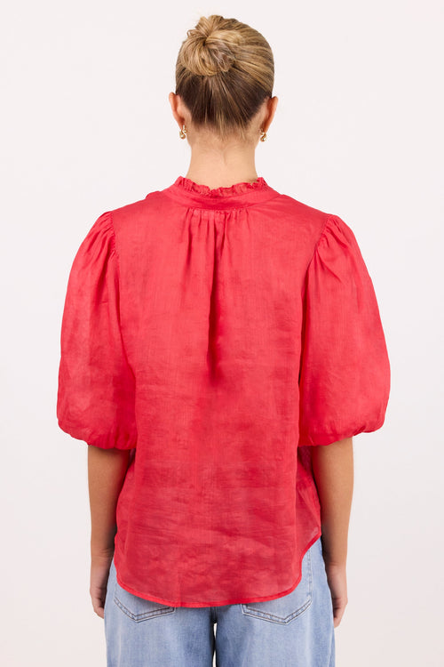 A woman viewed from behind wearing a red short puff-sleeve blouse and light blue jeans, standing against a plain white studio background.