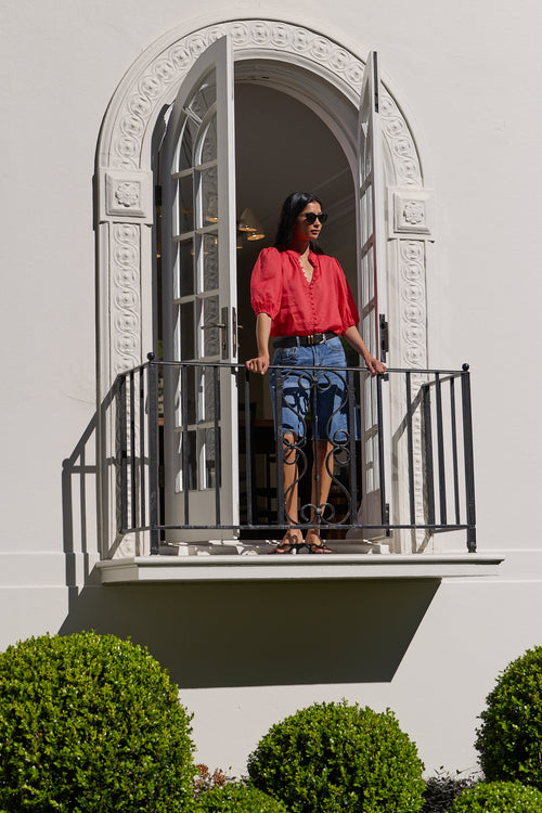 A woman wearing a bright coral-red blouse and distressed denim shorts stands on a white balcony with a black metal railing, set against a white stucco building facade with manicured green bushes below.