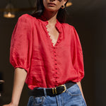 A woman wearing a vibrant red puff-sleeve blouse with a ruffled V-neckline and button front, paired with blue jeans and a black belt, standing in a doorway.