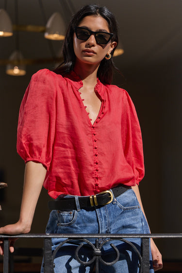 A woman wearing a vibrant red puff-sleeve blouse with a ruffled V-neckline and button front, paired with blue jeans and a black belt, standing in a doorway.