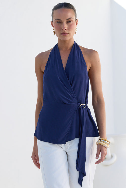 A woman modeling a navy blue halter wrap top with a side tie detail, paired with white pants, set against a bright white architectural background.