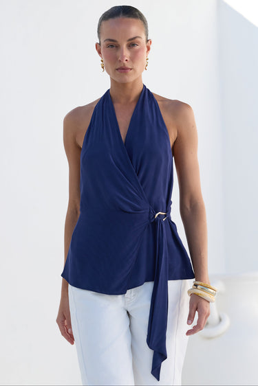 A woman modeling a navy blue halter wrap top with a side tie detail, paired with white pants, set against a bright white architectural background.