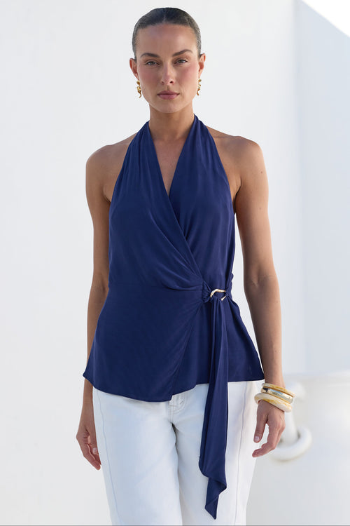A woman modeling a navy blue halter wrap top with a side tie detail, paired with white pants, set against a bright white architectural background.