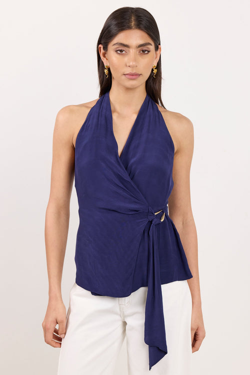 A woman modeling a navy blue halter wrap top with a side tie detail, paired with white pants, against a plain off-white studio background.