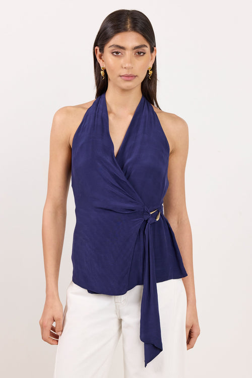 A woman modeling a navy blue halter wrap top with a side tie detail, paired with white pants, against a plain off-white studio background.
