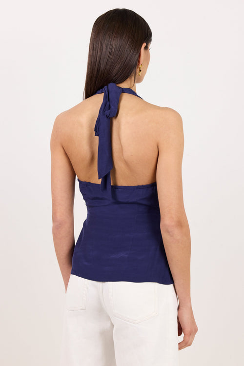 A woman shown from the back wearing a navy blue halter top with a tie at the neck and white pants, against a plain white studio background.